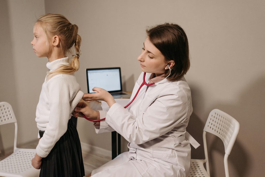 A female doctor in a white coat uses a stethoscope to examine a young girl's back during a medical checkup in an office.