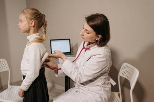 A female doctor in a white coat uses a stethoscope to examine a young girl's back during a medical checkup in an office.